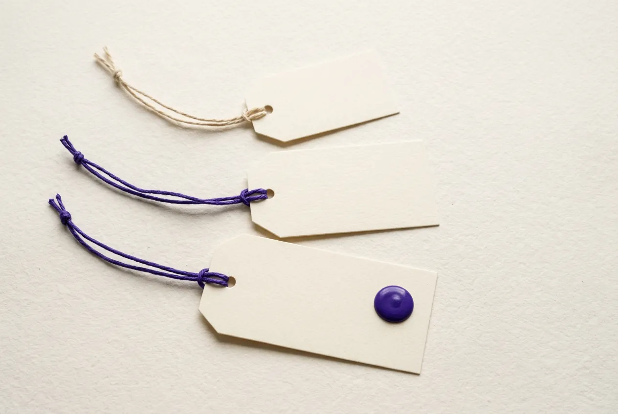 Three blank paper tags of growing size fanned out on cotton paper, two tied with violet string, the largest marked with a violet wax dot.